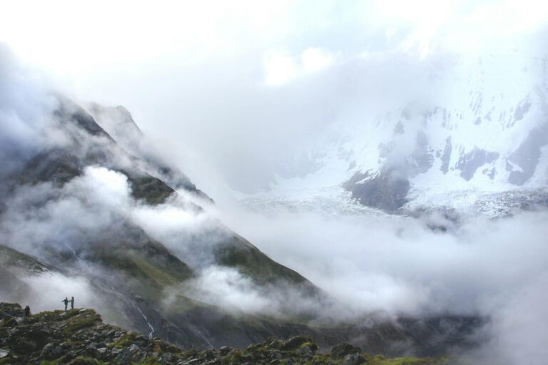 Trekkers on a rocky ridge below clouds swirling around Annapurna’s snowy face and glacier, Annapurna Base Camp Trek, Nepal